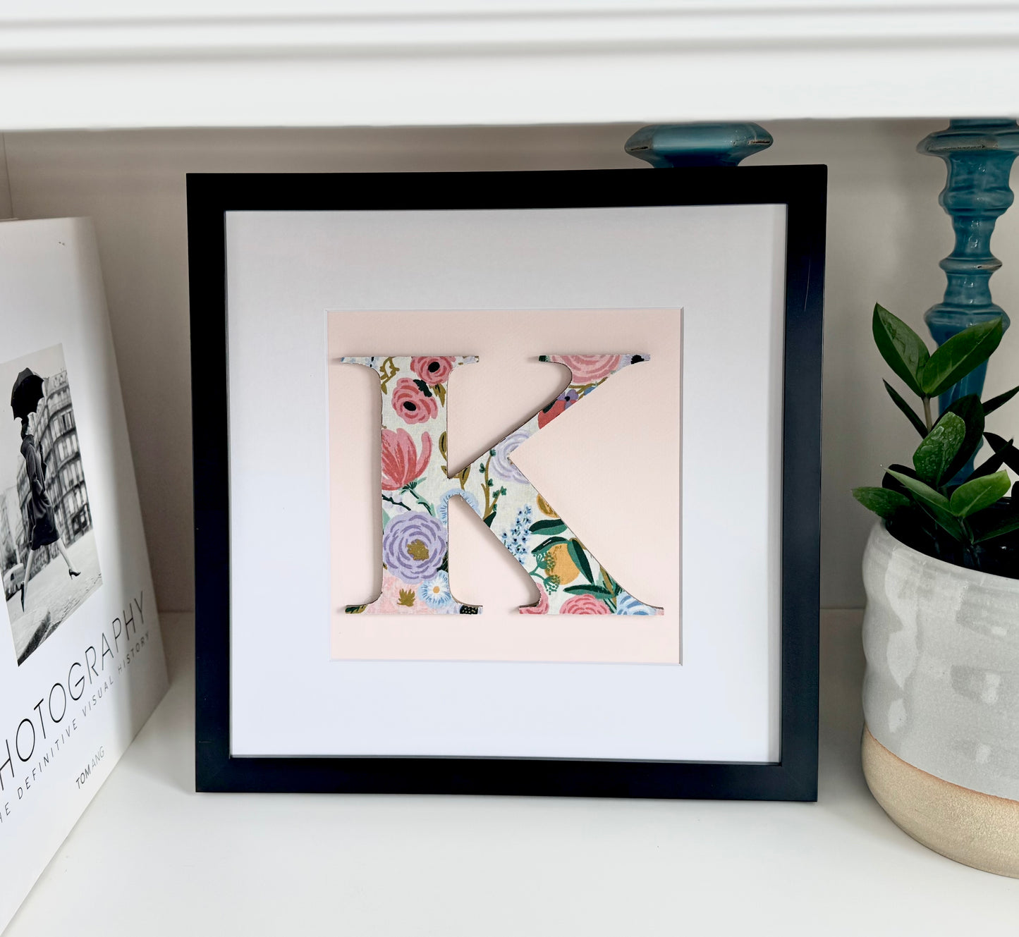 Floral Letter K on a pink background in a black frame, sitting on a shelf