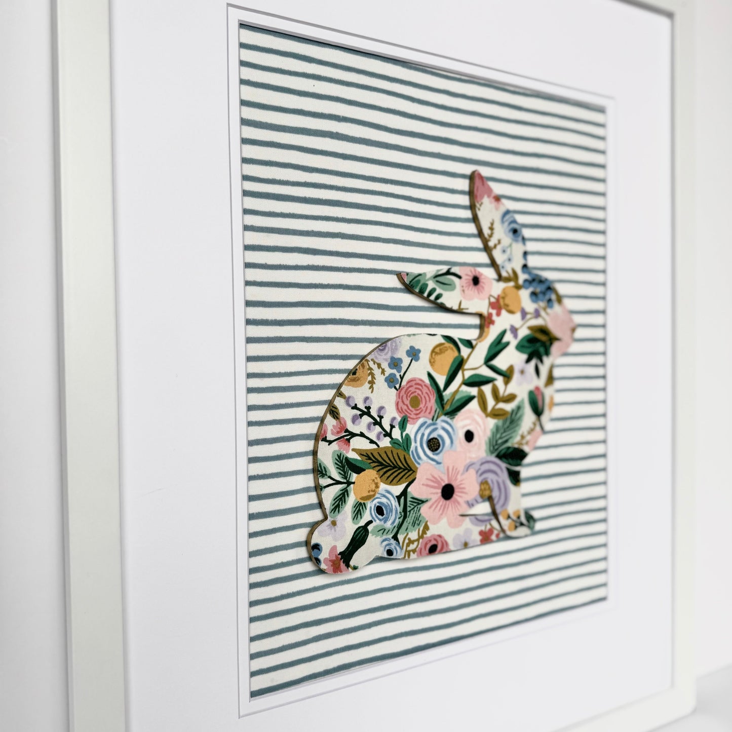 White, framed Rifle Paper Co. Floral bunny on a blue striped background, hanging on a wall