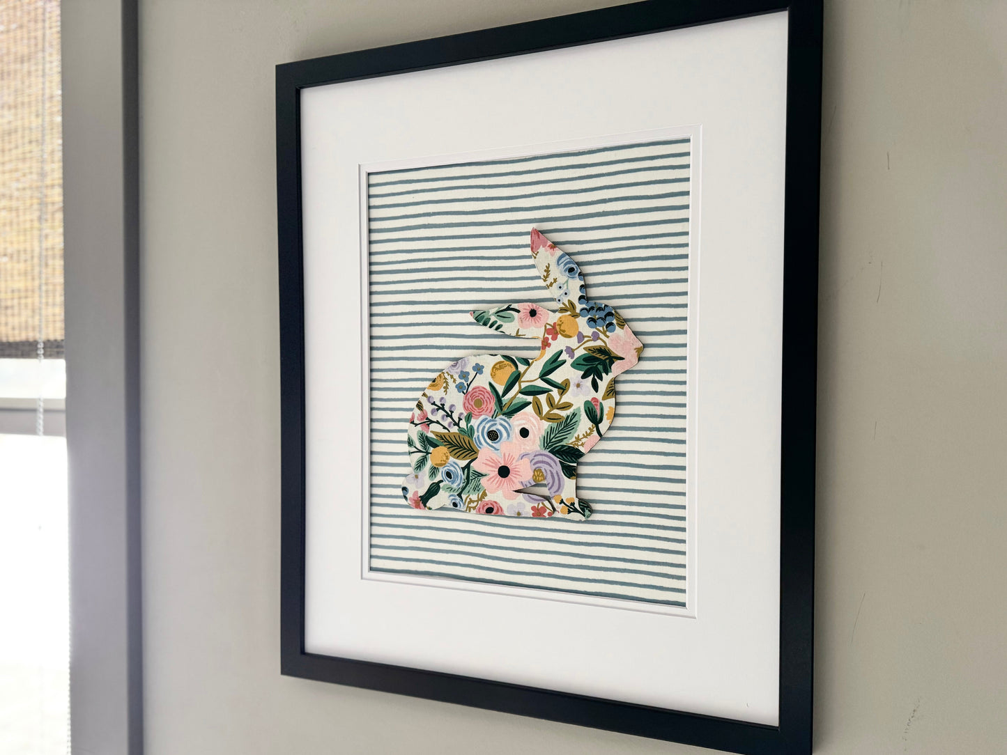 Black, framed Rifle Paper Co. Floral bunny on a blue striped background, hanging on a wall