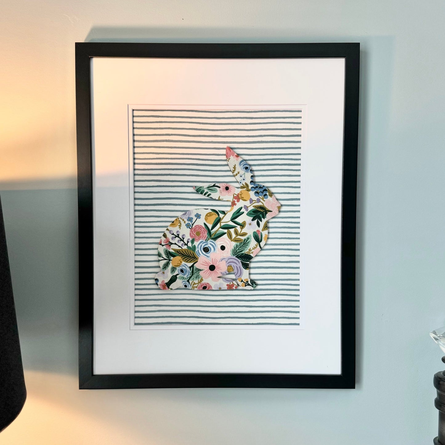 Black, framed Rifle Paper Co. Floral bunny on a blue striped background, hanging on a wall