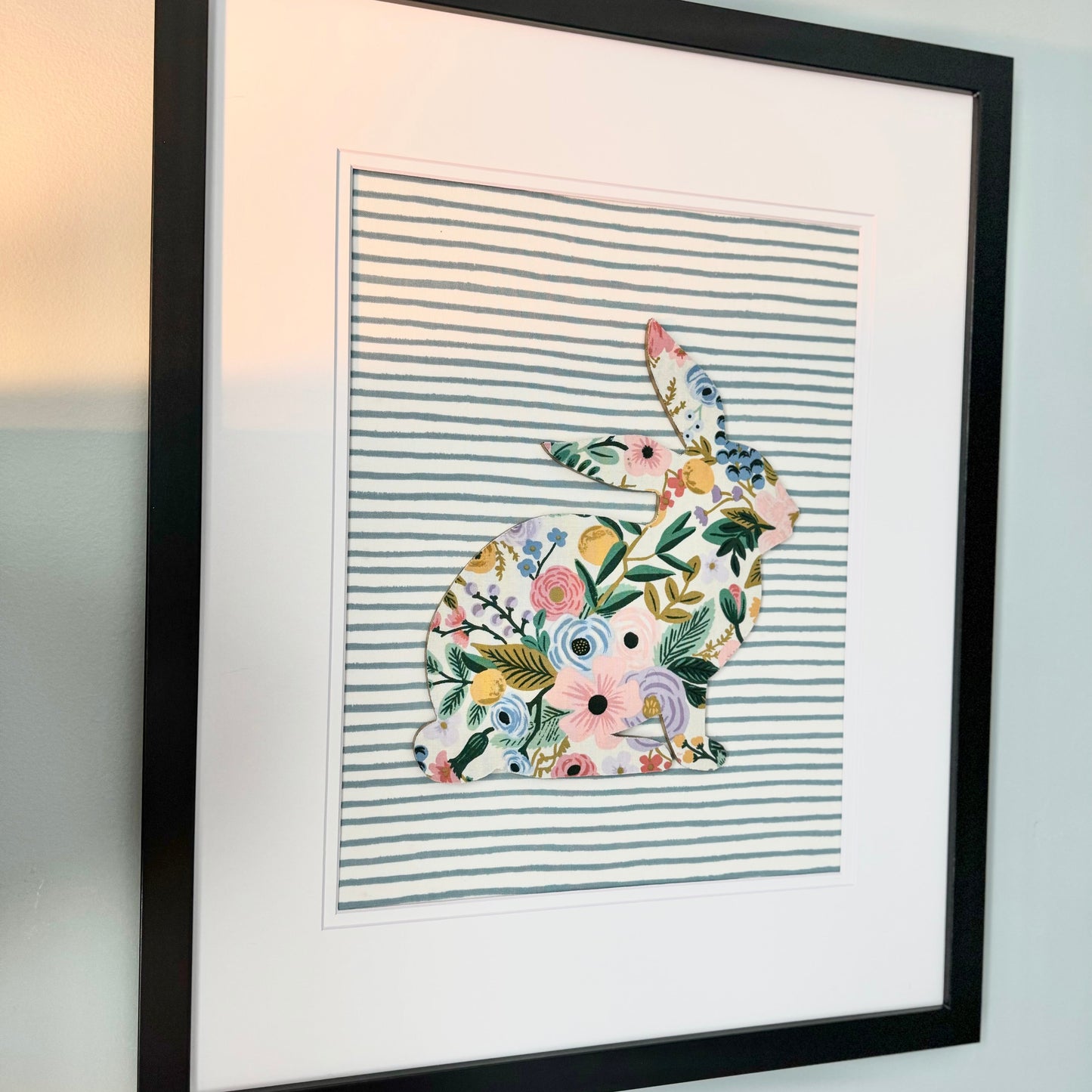 Black, framed Rifle Paper Co. Floral bunny on a blue striped background, hanging on a wall