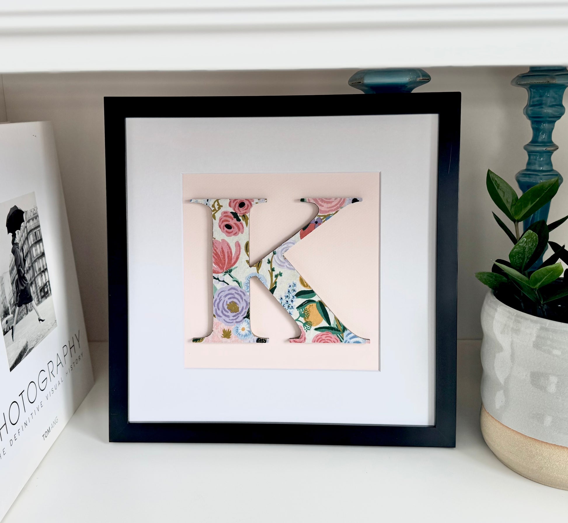Floral Letter K on a pink background in a black frame, sitting on a shelf
