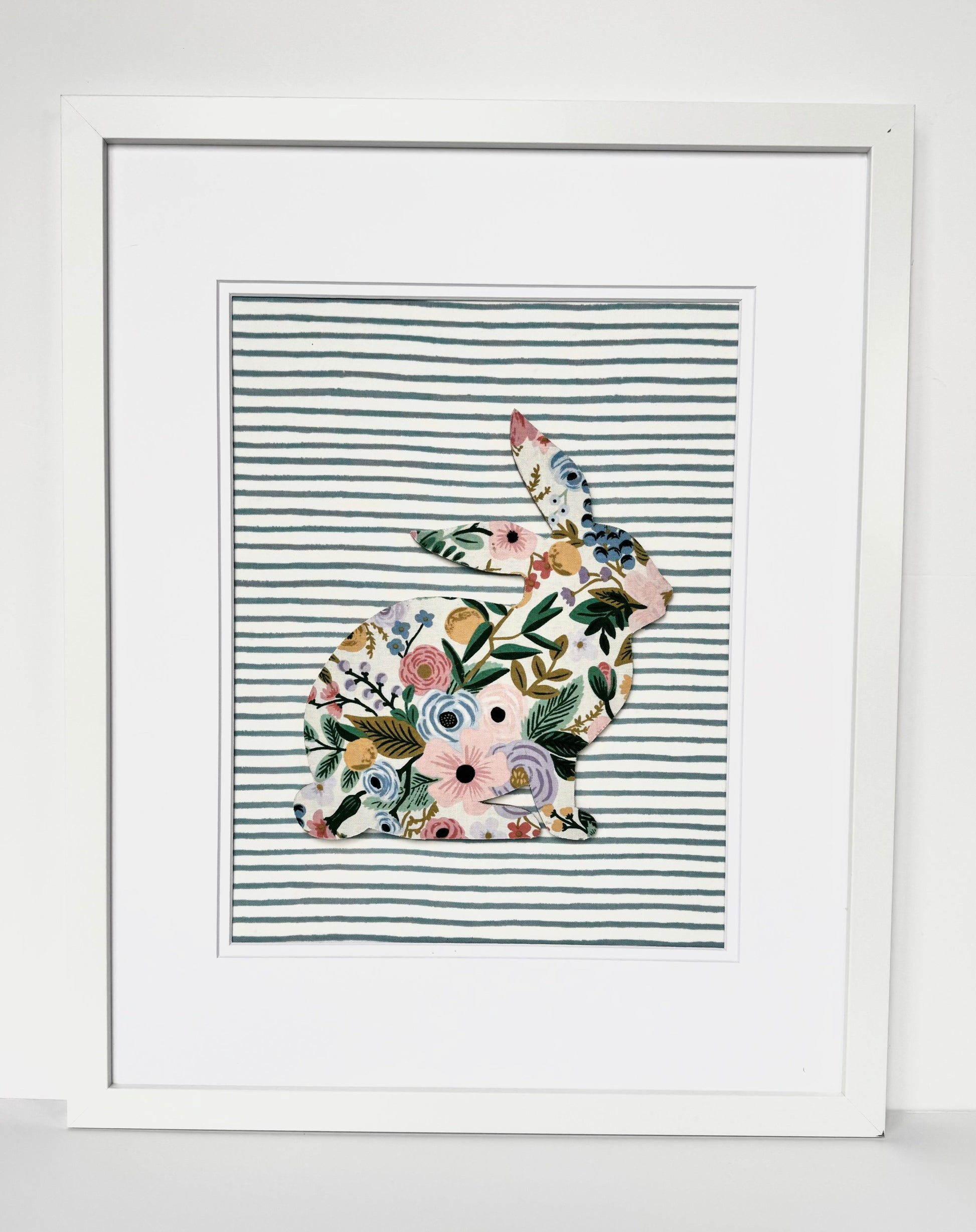 White, framed Rifle Paper Co. Floral bunny on a blue striped background, hanging on a wall