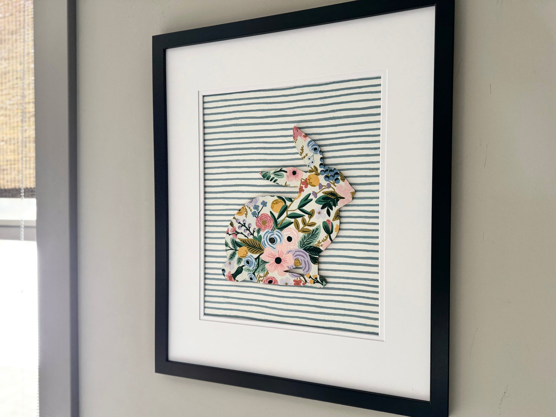 Black, framed Rifle Paper Co. Floral bunny on a blue striped background, hanging on a wall